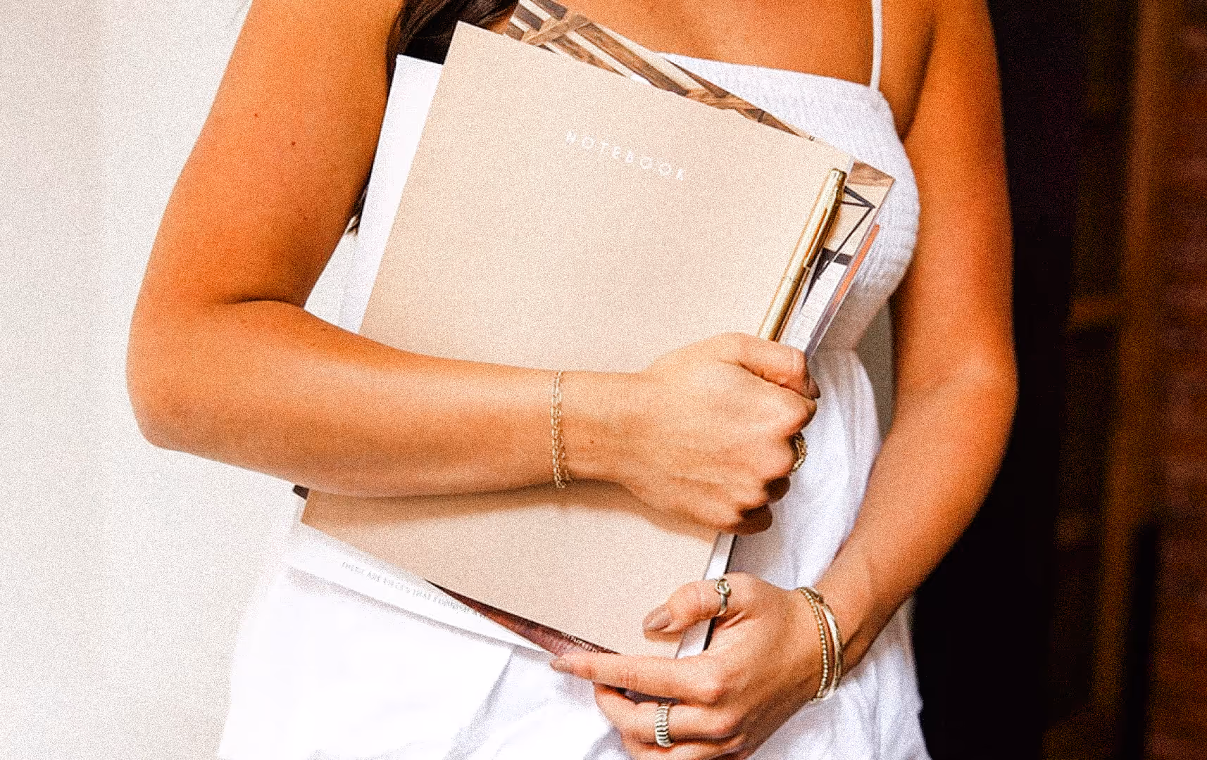A person in a white dress holds a beige notebook and a pen, with gold jewelry on their wrist and fingers. The scene conveys a calm, organized vibe.