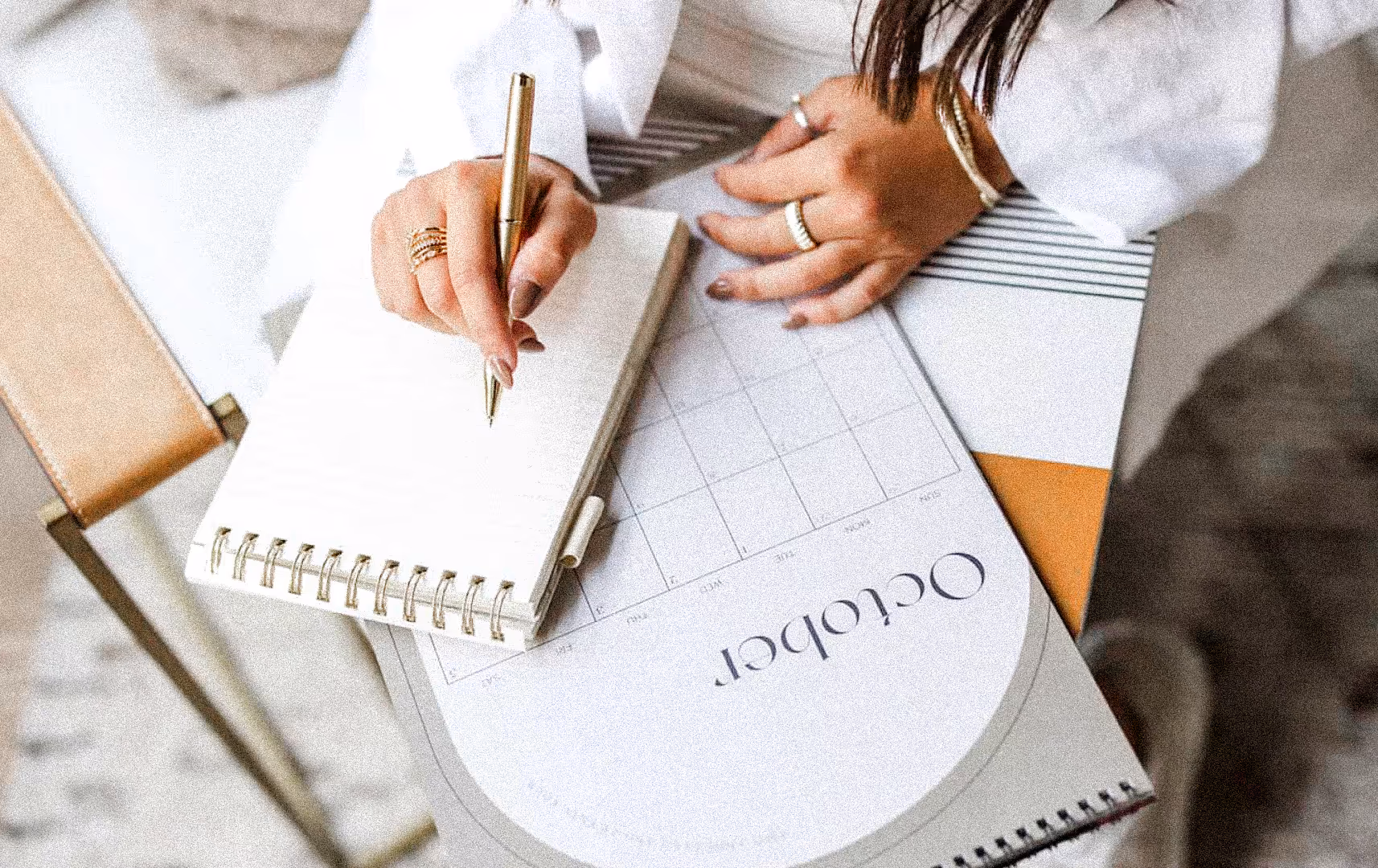 A person in white writes in a spiral notebook on a desk with an October calendar. The scene is calm, organized, and focused, conveying productivity.