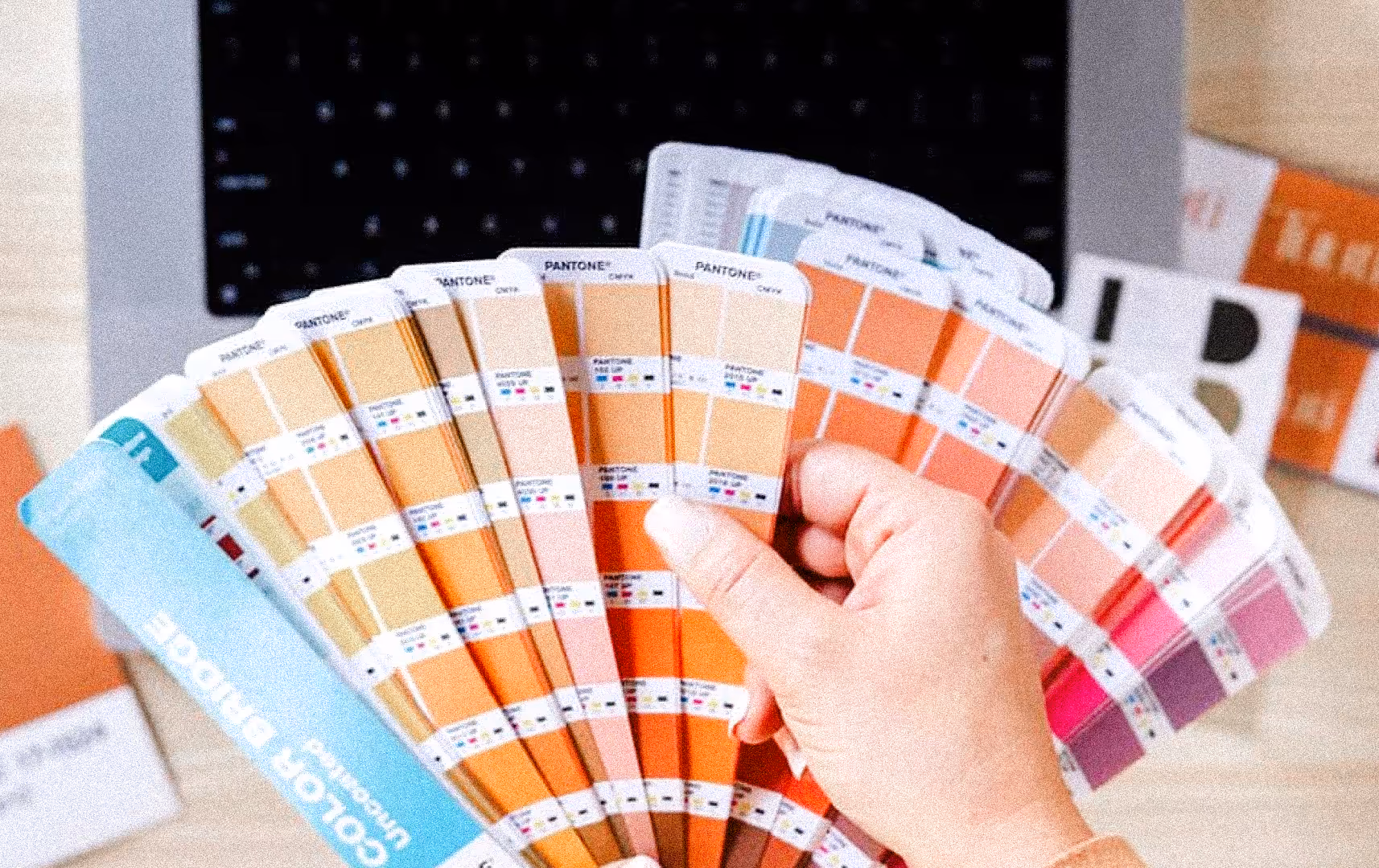 A person holds a fanned-out Pantone color guide showing shades of orange, pink, and yellow over a laptop, suggesting design and creativity.