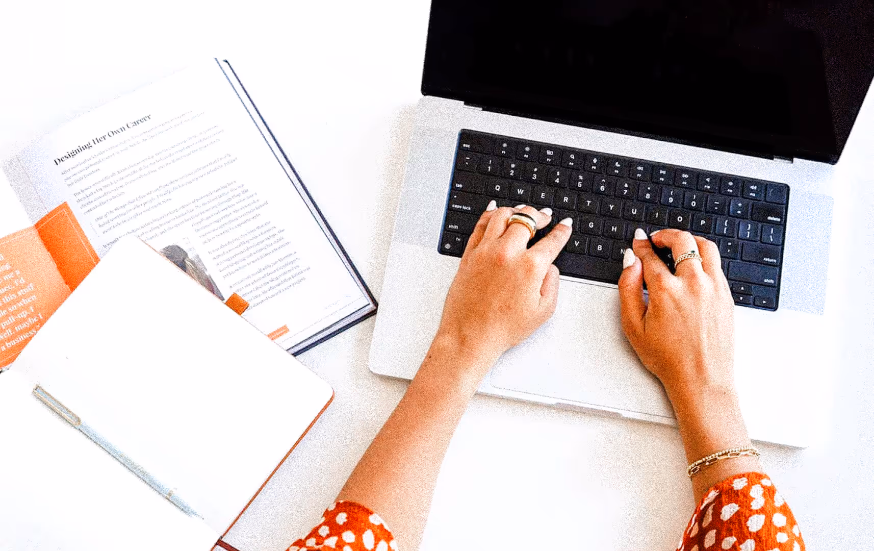 Hands typing on a laptop keyboard beside an open notebook and book on a white desk. The setting is bright and conveys a focused work atmosphere.