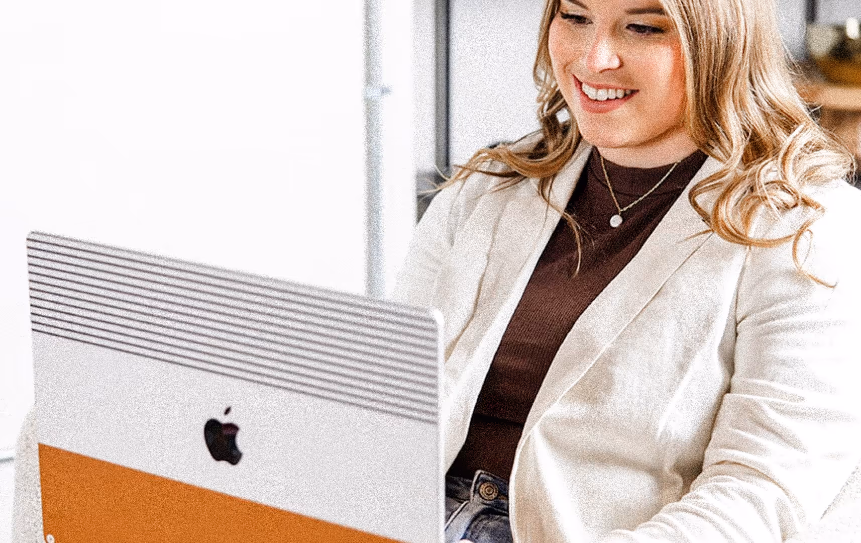A woman with long hair sits at a desk smiling while using a laptop. She wears a beige blazer and a dark top, creating a relaxed and professional atmosphere.