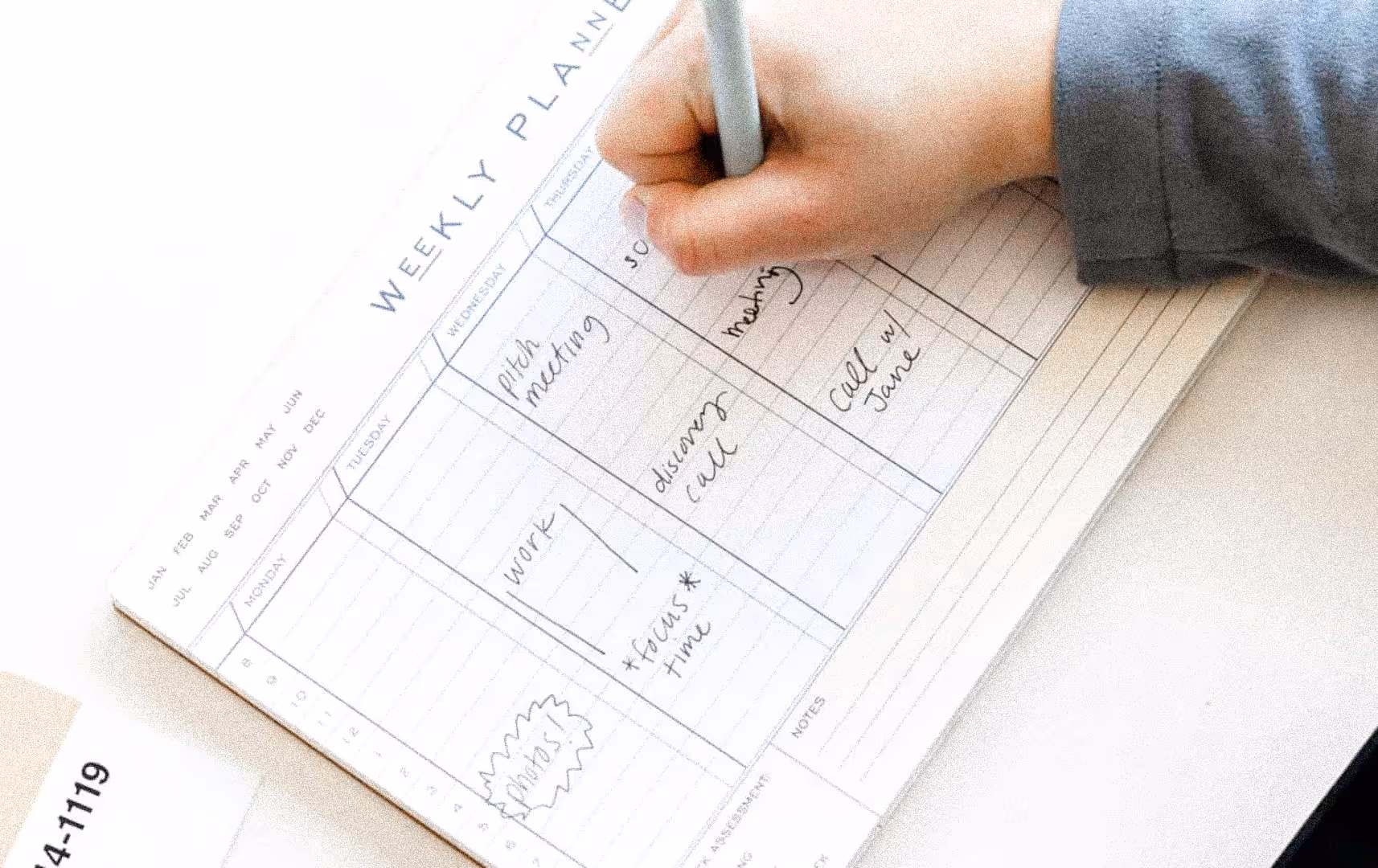 A hand holding a pen writes on a weekly planner. The planner includes notes like "pitch meeting" and "focus time," conveying organization and productivity.