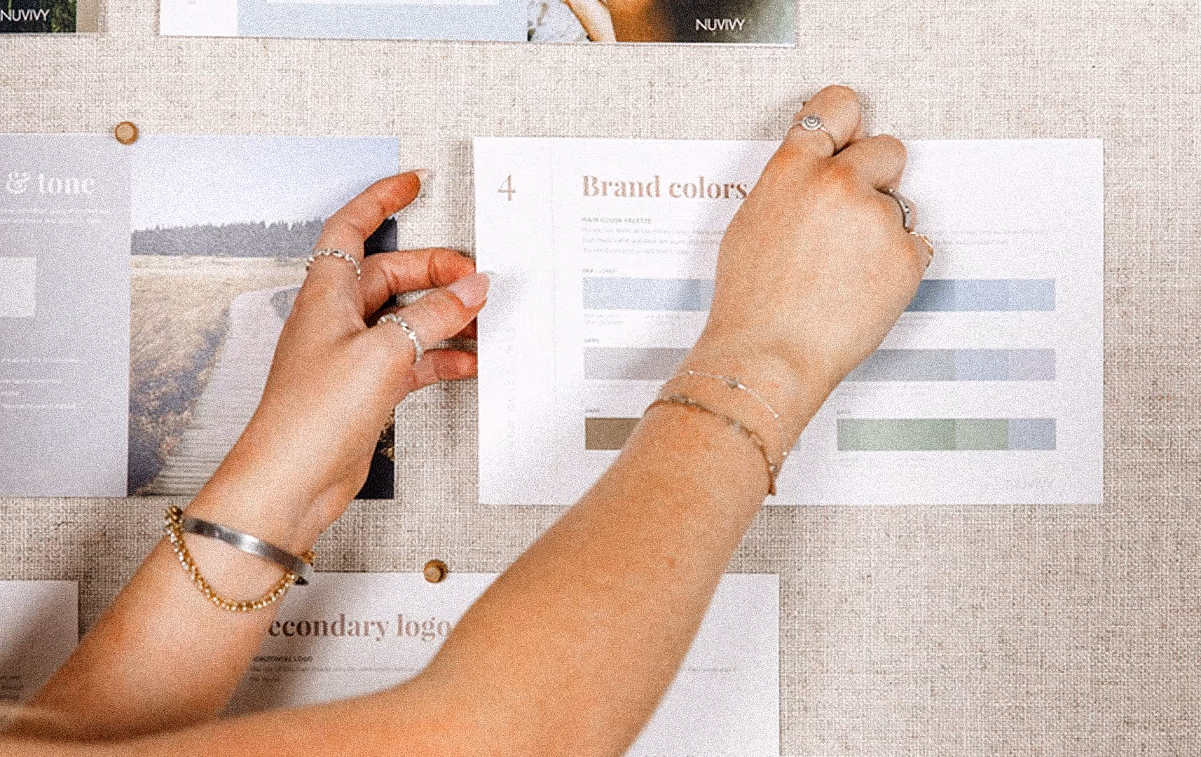 Two hands pin a brand colors chart on a beige corkboard, surrounded by design notes. The scene conveys creativity and organization.