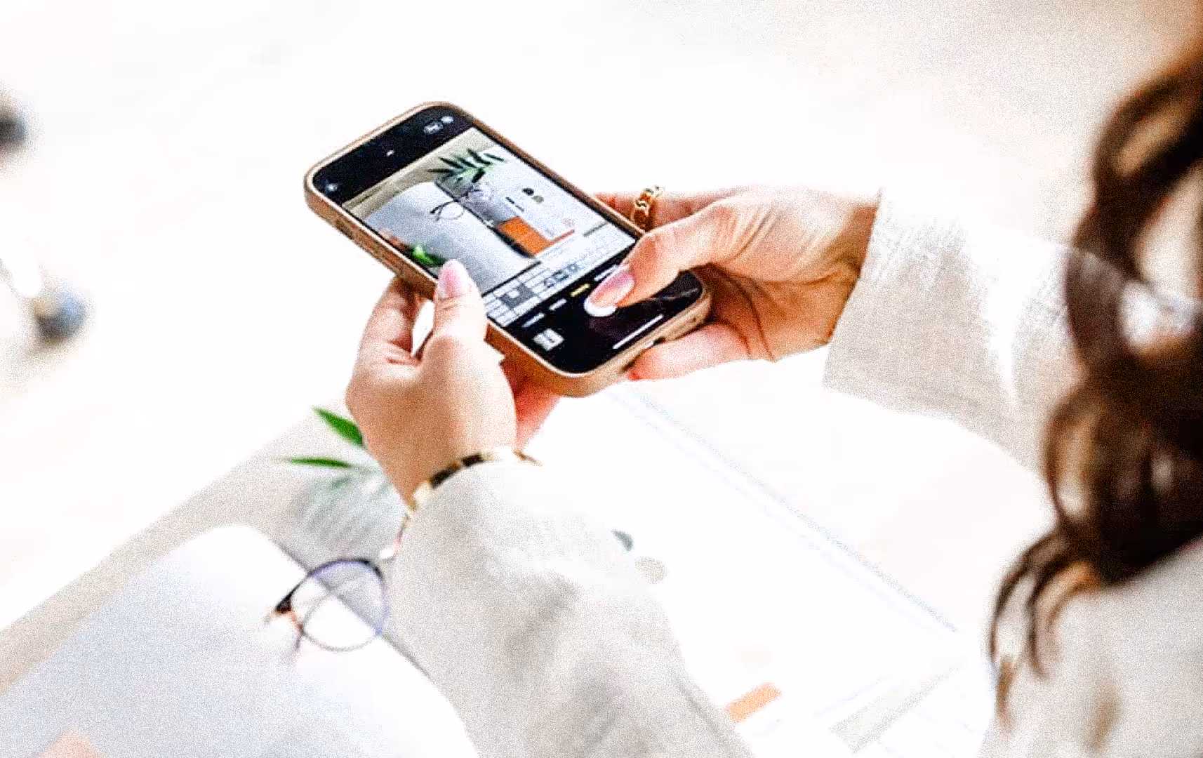 A person in white clothing holds a smartphone, photographing a desk with glasses, a notebook, and a small plant, conveying a focused, creative mood.