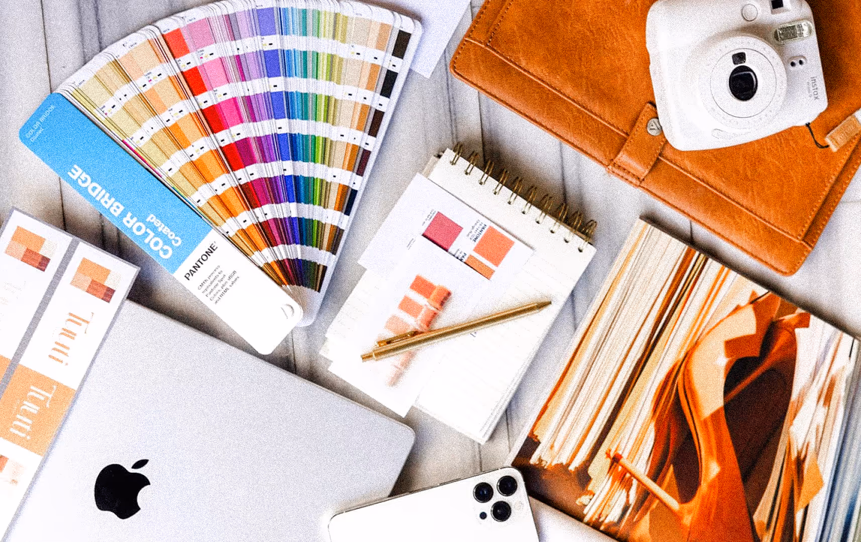 A creative workspace with a colorful Pantone swatch book, a notebook with a pen, an open laptop, a smartphone, and a camera, conveying inspiration.