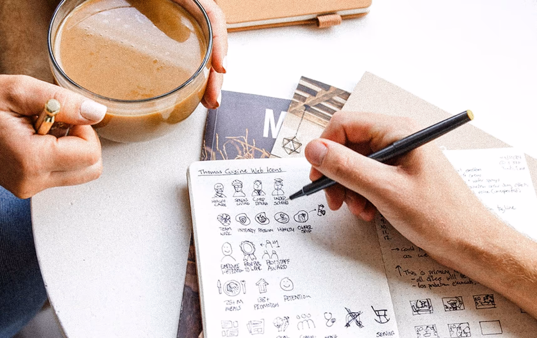 Two people are at a table. One holds a coffee cup, while the other sketches ideas in a notebook. The scene feels creative and collaborative.
