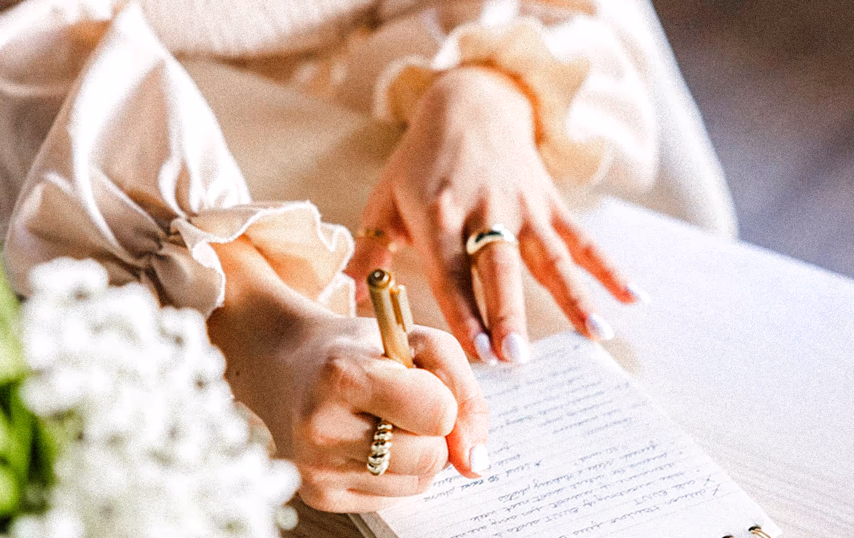 A woman in a ruffled blouse writes in a notebook with a gold pen. Her hands display elegant rings. Nearby, blurred white flowers add a soft touch.