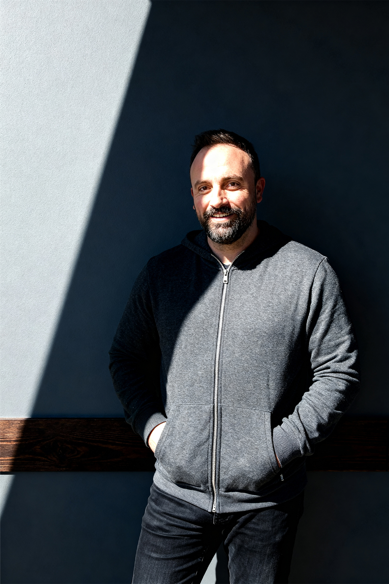 A man with a beard stands smiling in partial shadow against a dark wall, wearing a gray zip-up hoodie. Sunlight creates a dramatic diagonal pattern.