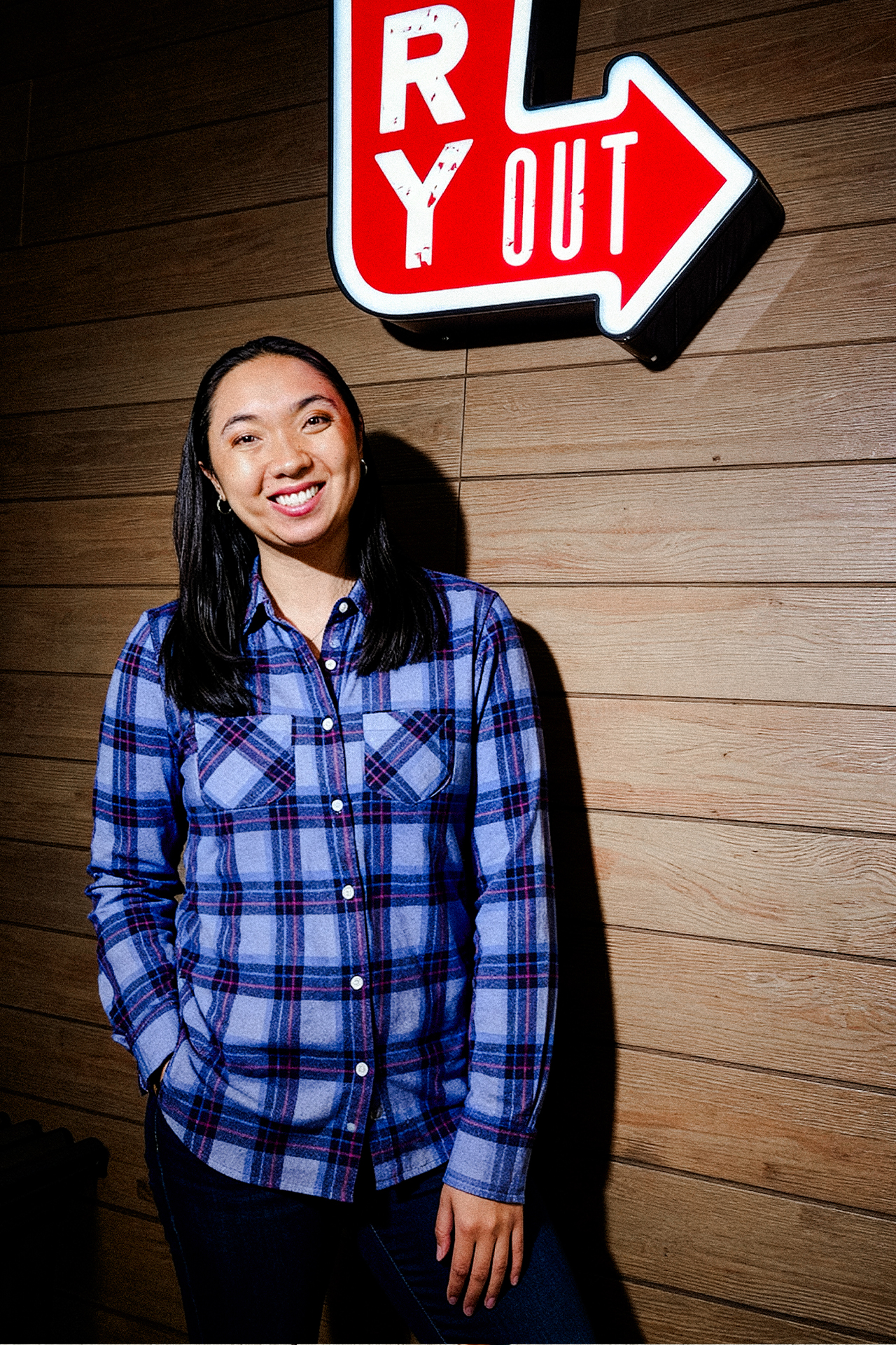 Smiling person in a blue plaid shirt stands against a wooden wall beneath an illuminated red and white "OUT" arrow sign. Warm, inviting atmosphere.