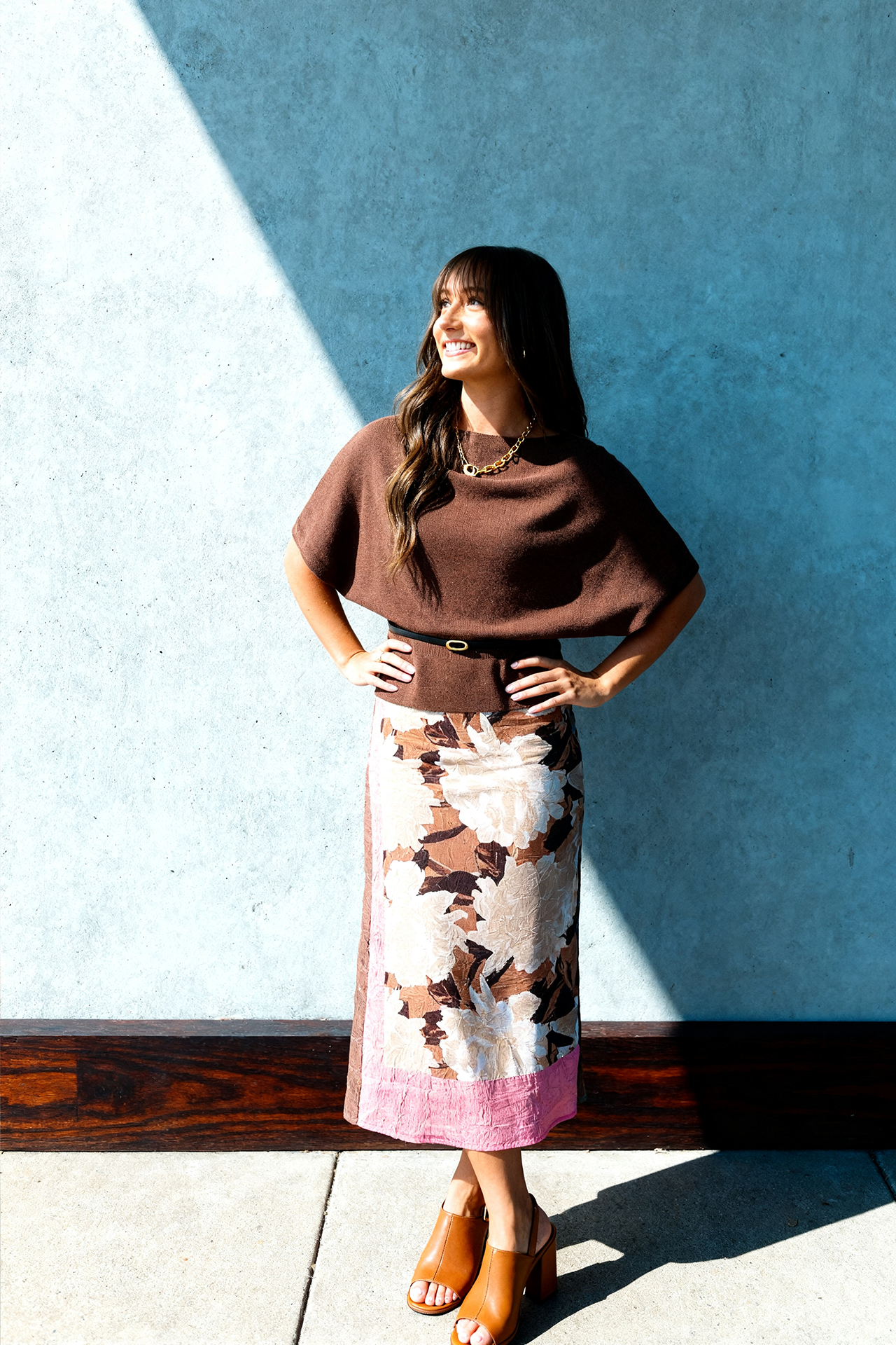 A woman stands confidently against a sunlit, blue wall. She wears a brown top, floral skirt, and tan heels, smiling and looking to the side.