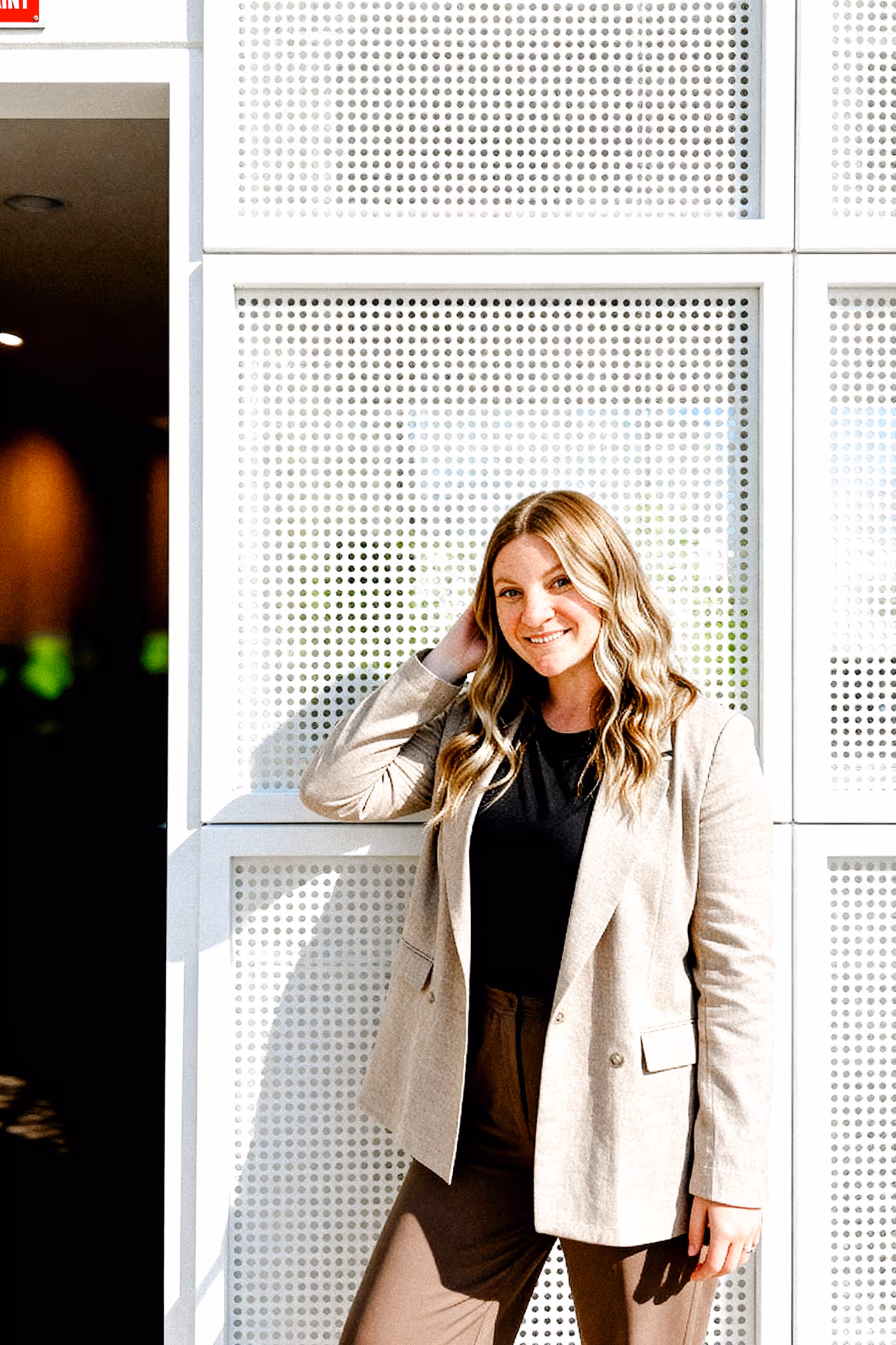 A woman with wavy hair smiles warmly, leaning against a white, perforated wall. She wears a beige blazer and dark top, exuding confidence and warmth.
