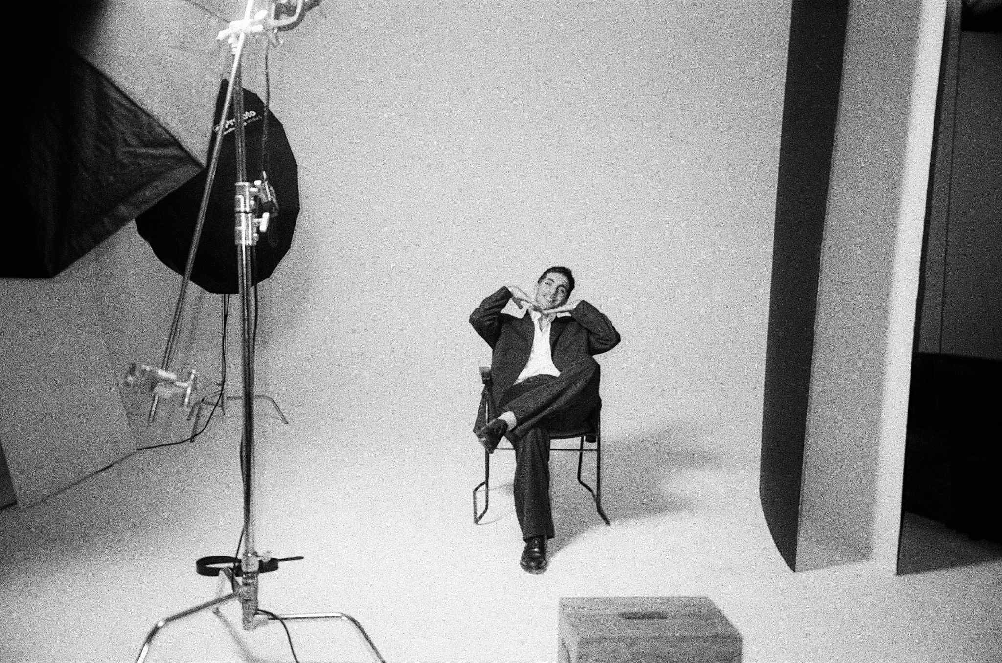 Un homme en tailleur sombre assis sur une chaise dans un studio photo, posant avec les mains sous le menton, entouré d'équipements d'éclairage.