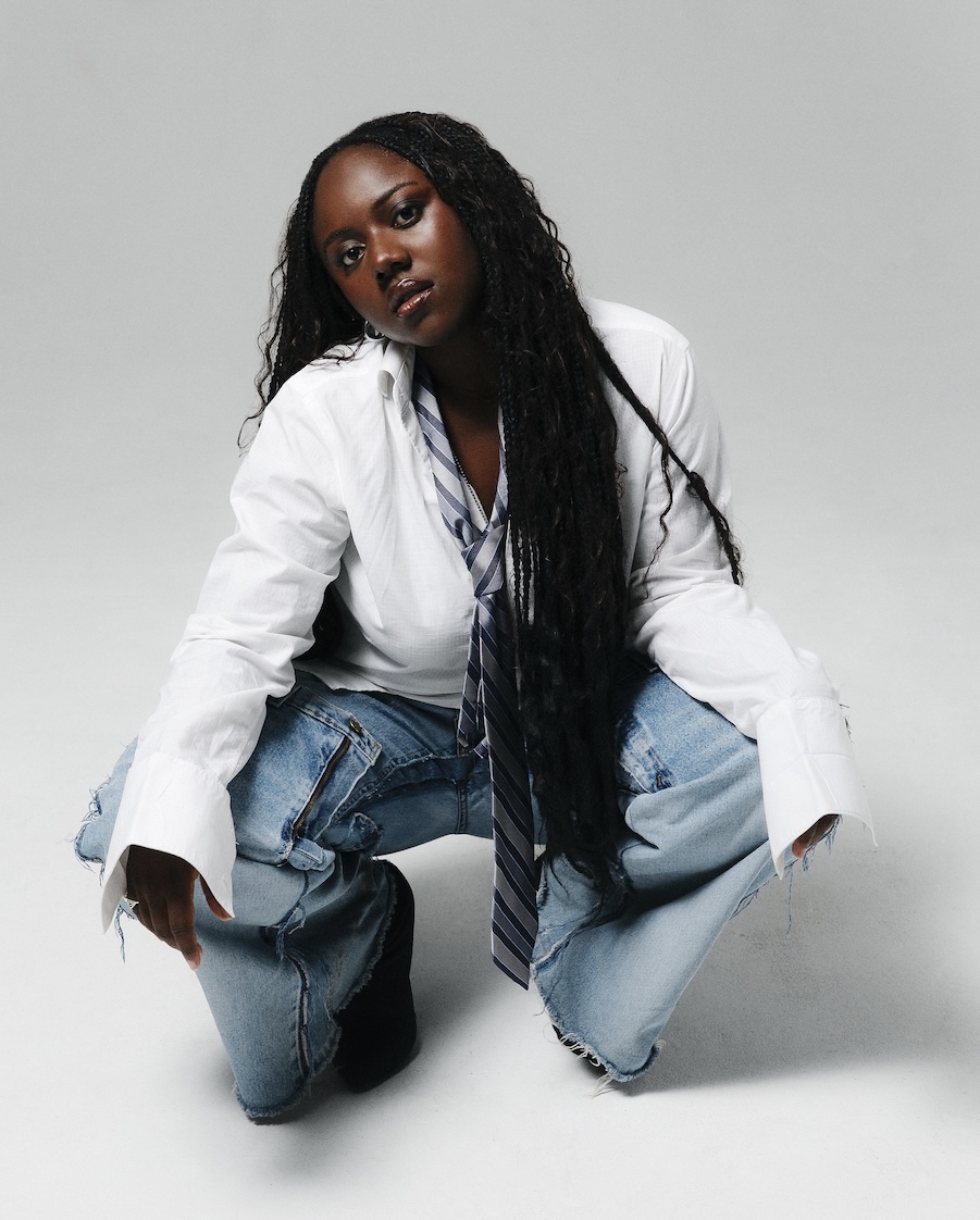 Femme noire aux longs cheveux tressés, accroupie, portant une chemise blanche, une cravate rayée et un jean déchiré.