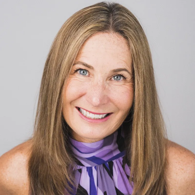 Smiling middle-aged woman with long straight blonde hair wearing a purple patterned top against a light gray background.