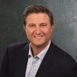 Smiling man with dark hair wearing a white shirt and dark blazer against a blurred dark background.