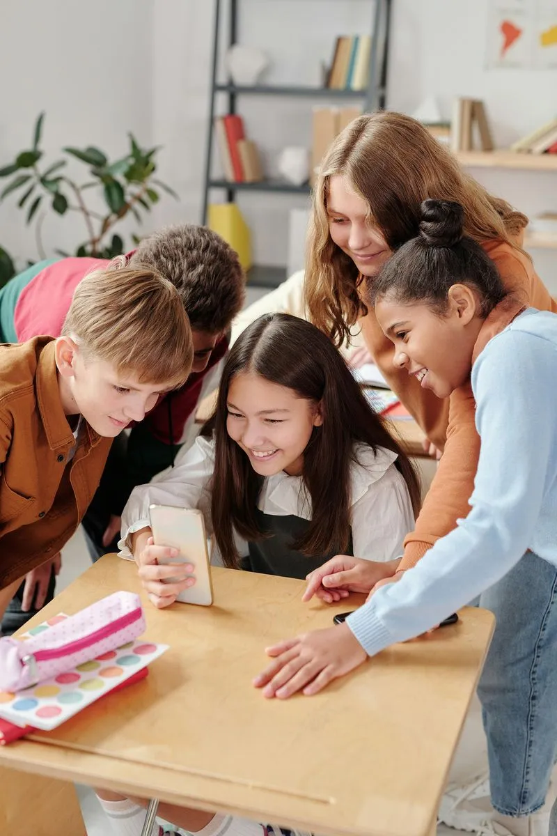 A group of children gathered around a schoolgirl's desk, looking at her phone