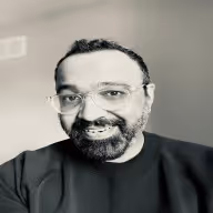 Smiling man with glasses, beard, and curly hair wearing a black shirt against a plain background.