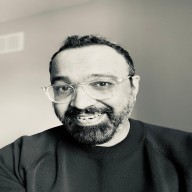 Smiling man with glasses, beard, and curly hair wearing a black shirt against a plain background.