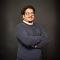 Man with glasses and mustache smiling, wearing a dark sweater over a white collared shirt, standing with arms crossed against a dark background.