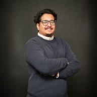 Man with glasses and mustache smiling, wearing a dark sweater over a white collared shirt, standing with arms crossed against a dark background.