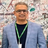 Smiling man wearing glasses and a gray blazer over a dark shirt, standing in front of a wall covered with colorful graffiti and signatures.