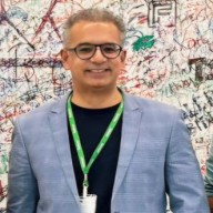 Smiling man wearing glasses and a gray blazer over a dark shirt, standing in front of a wall covered with colorful graffiti and signatures.