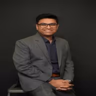 Smiling man wearing glasses, a gray suit, and a dark shirt sitting against a black background.