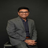 Smiling man wearing glasses, a gray suit, and a dark shirt sitting against a black background.