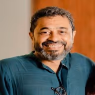 Smiling man with short curly hair and beard wearing a dark green shirt against a blurred warm background.