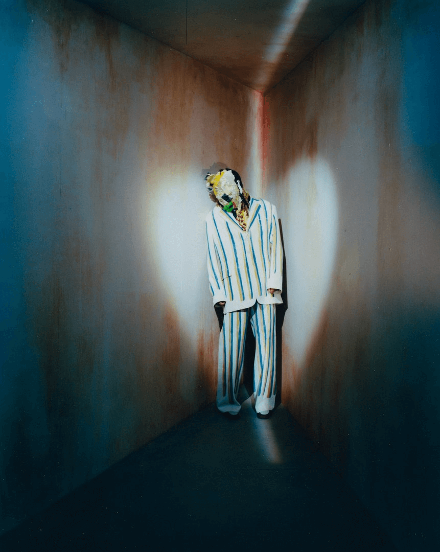 Masked figure in white suit with red scarf stands in surreal room lit by strong light beam.