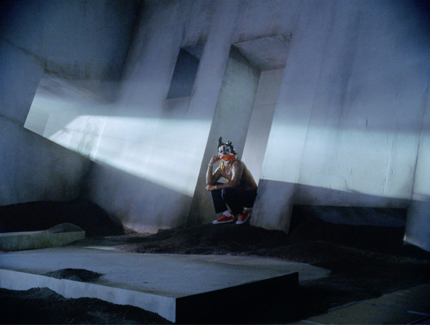 Masked figure in white suit with red scarf stands in surreal room lit by strong light beam.