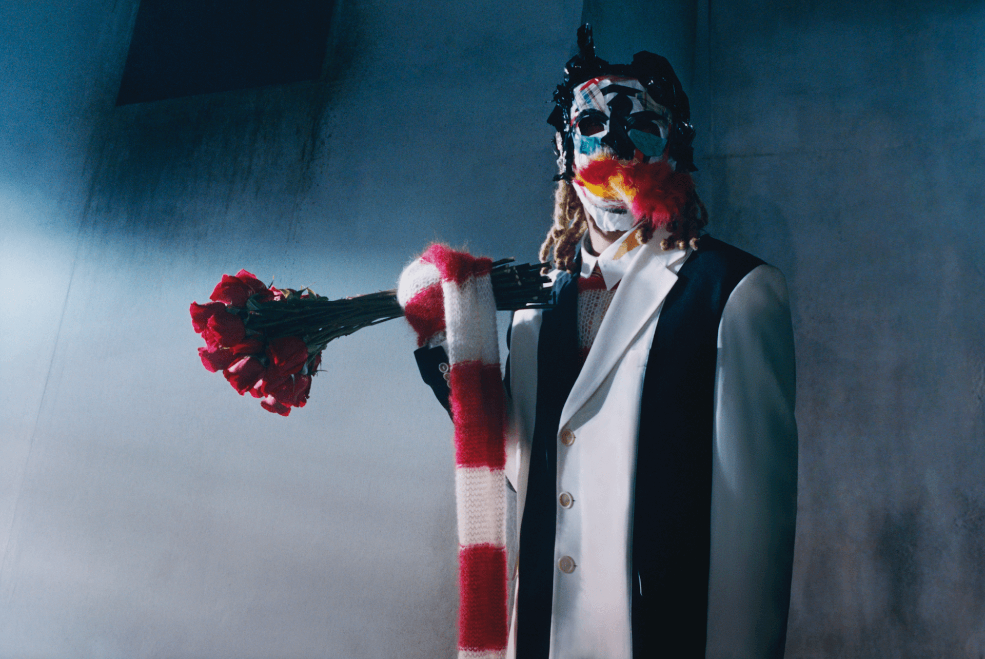 Masked figure in white suit with red scarf holding bouquet of red roses in dim setting.