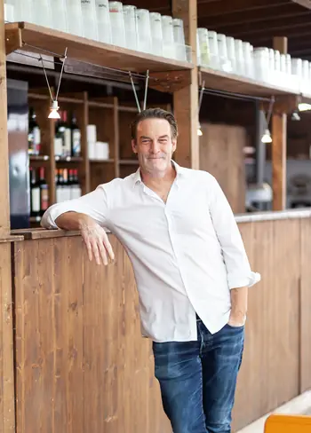 Mann lehnt lächelnd an einer Holzbar in einem rustikalen Ambiente mit Regalen voller Gläser im Hintergrund.