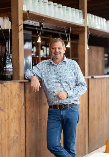 Ein lächelnder Mann lehnt an einer hölzernen Bar und hält ein Glas Getränk in der Hand.