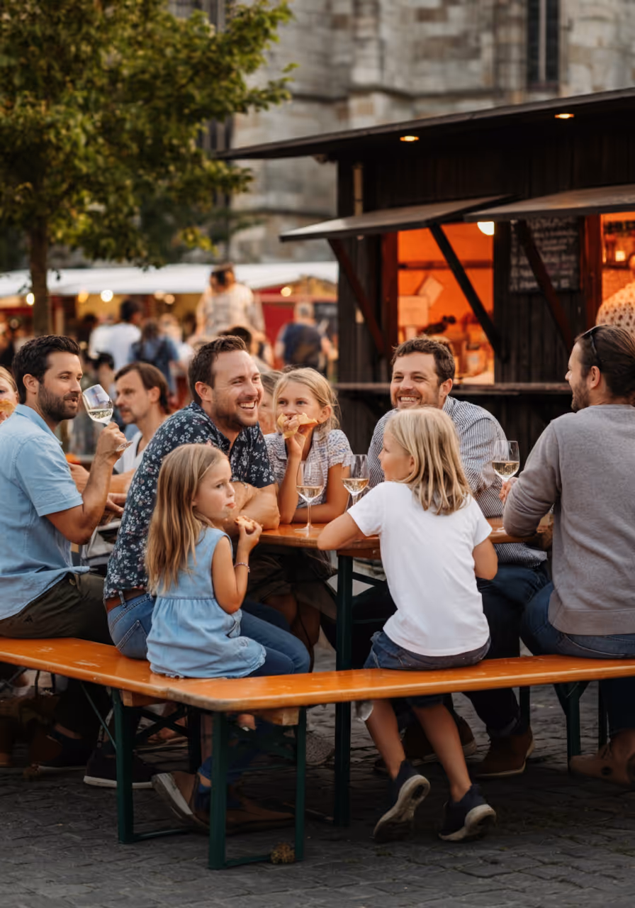 Eine Gruppe von Erwachsenen und Kindern sitzt an einem Biergartentisch im Freien und genießt Essen und Wein.
