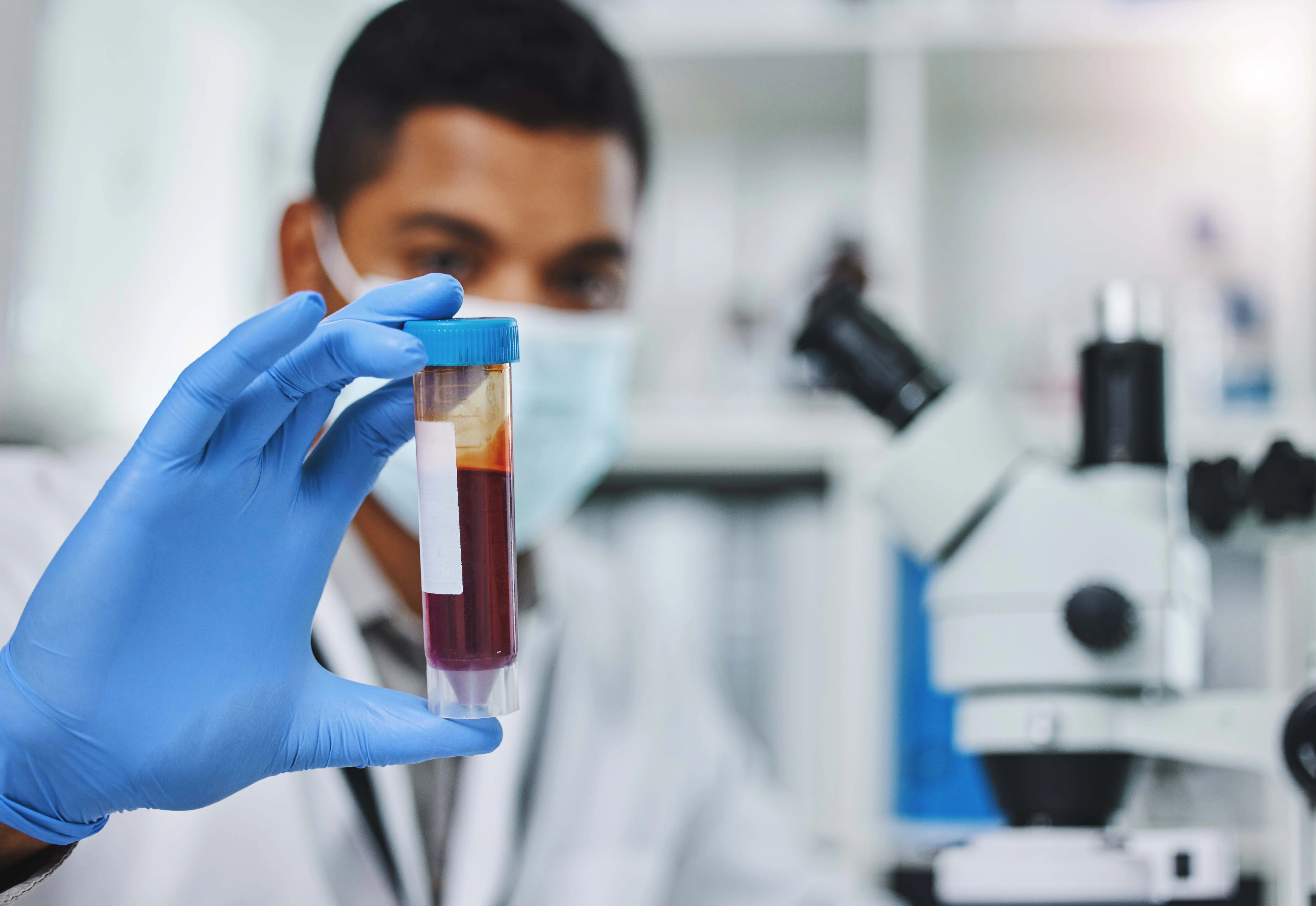 A lab worker wearing blue gloves and a mask holds a test tube with a blood sample for analysis.