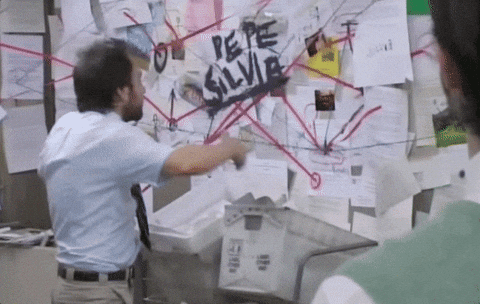 Animated GIF showing a man in an office gesturing wildly at a wall covered in papers, photos, and red string connecting them, as he tries to explain a chaotic web of connections.