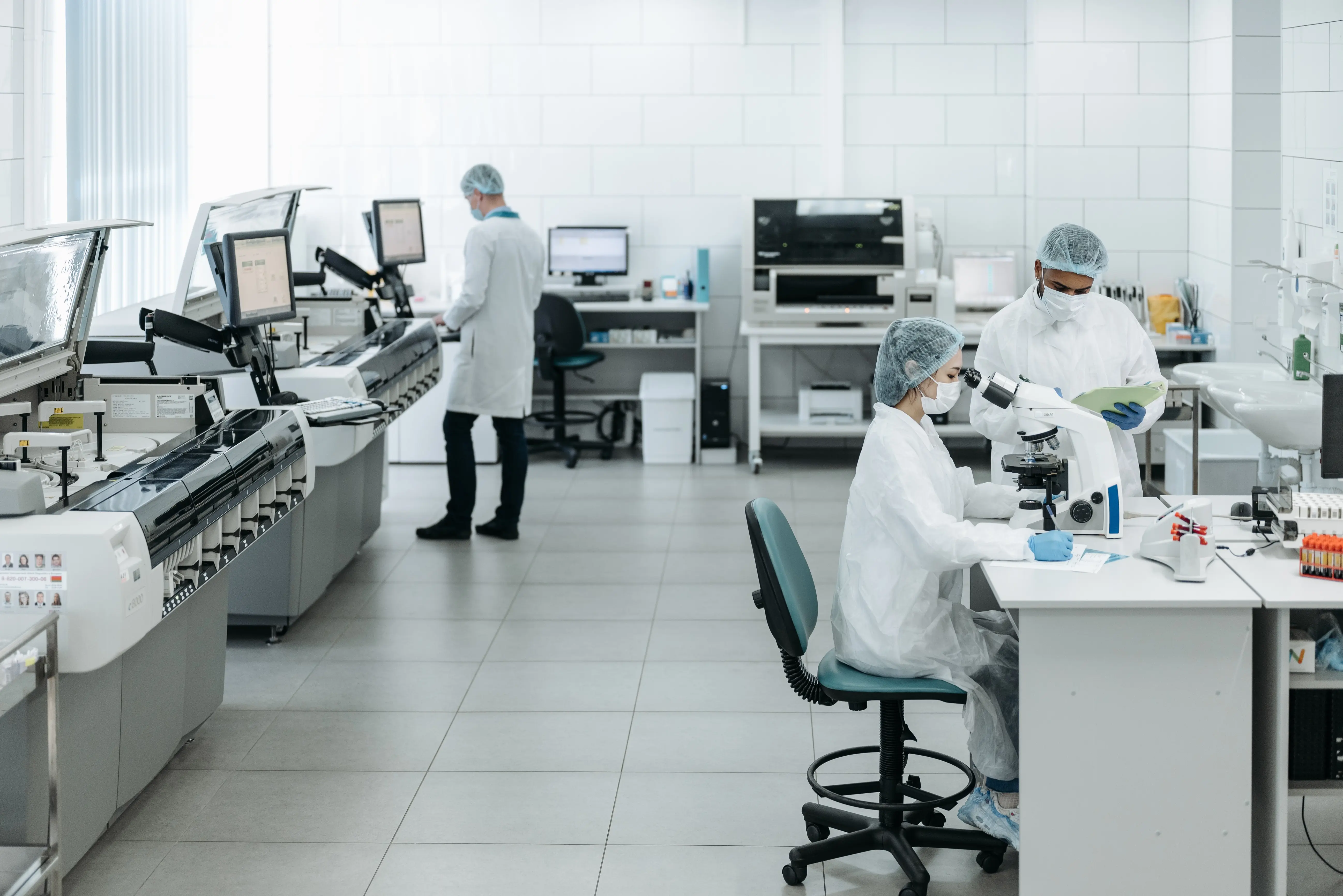 A laboratory scientist wearing a face mask and blue gloves examining a blood sample in a laboratory. A microscope and lab equipment are visible in the blurred background
