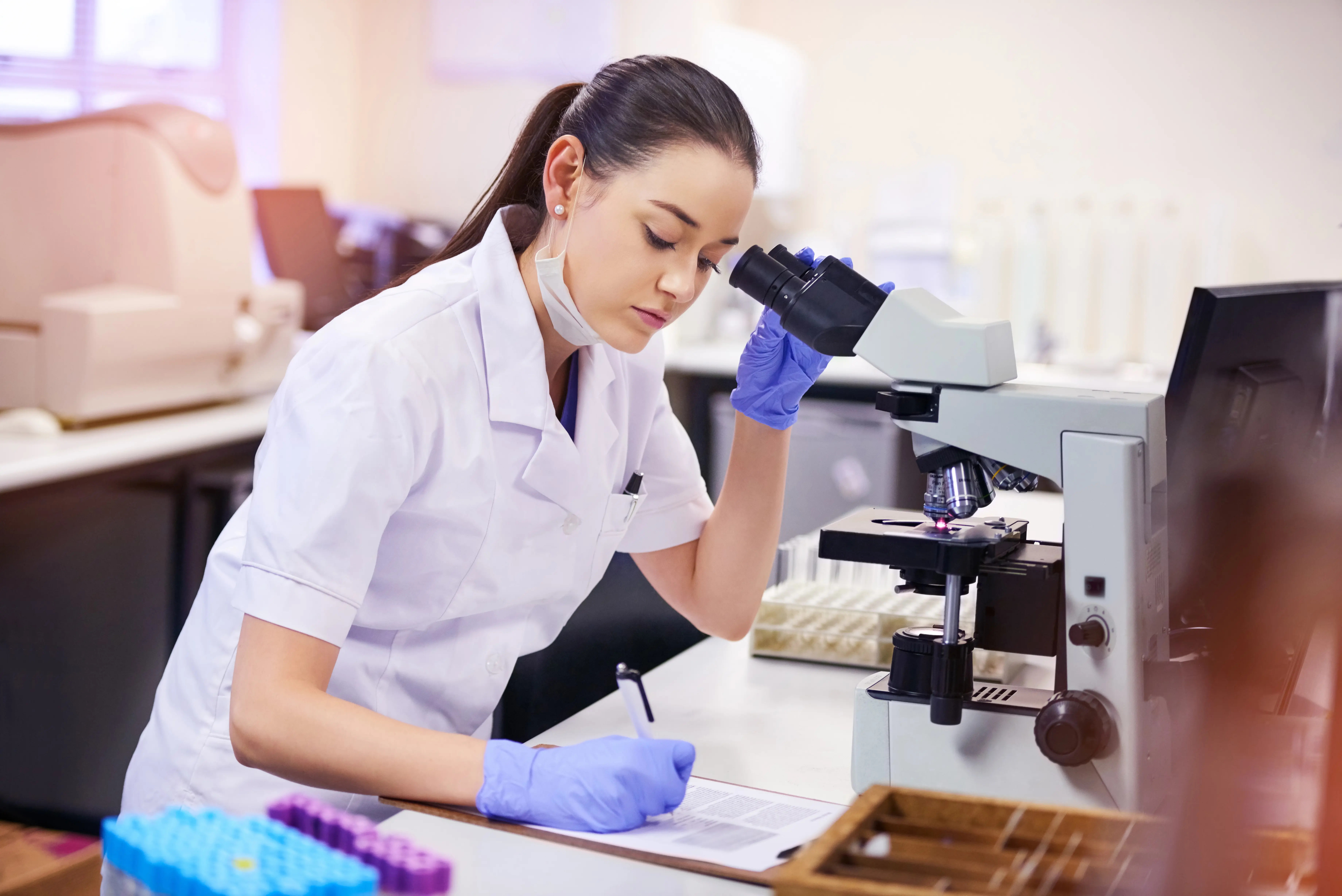 Female scientist in a white lab coat and blue gloves using a microscope in a research laboratory.