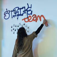 Person with long hair spray painting the words 'strike team' on a white wall above a grid of small photos.