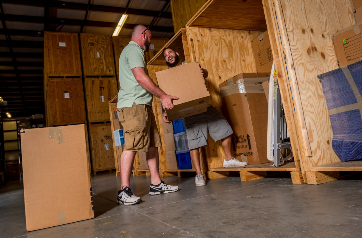 Two professional movers working together to load cardboard boxes into a wooden storage vault in a warehouse