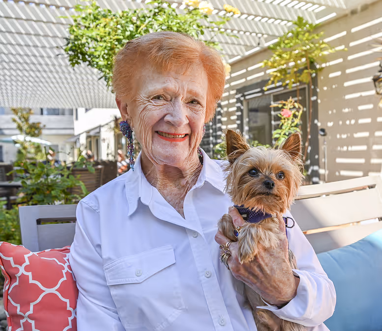 Senior woman with short red hair smiles warmly while holding a small Yorkshire Terrier. They're outdoors on a sunny day, with buildings and greenery in the background.