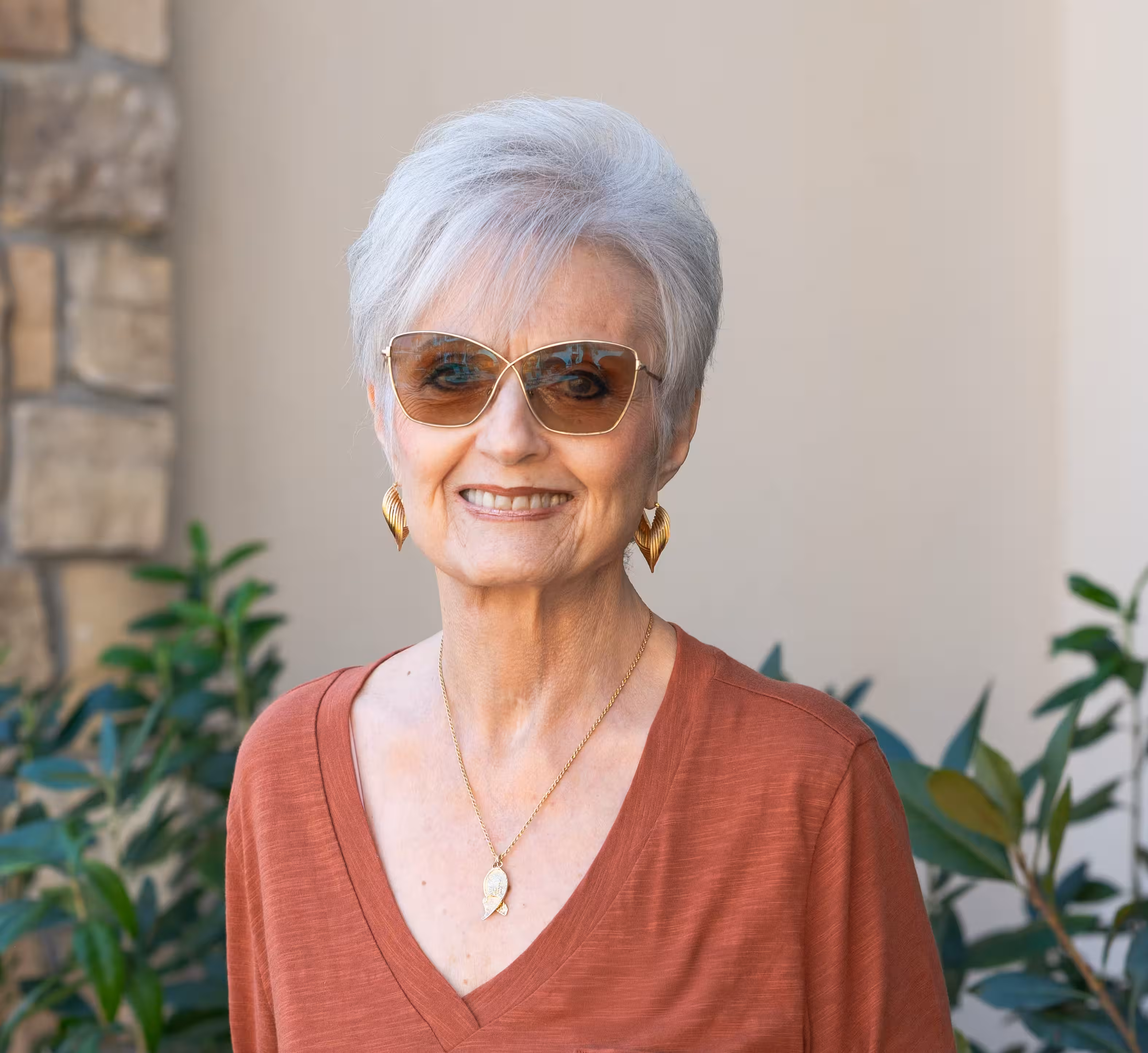 Elderly woman with short white hair smiles warmly, wearing sunglasses and gold earrings. She is dressed in a rust-colored top with plants in the background.