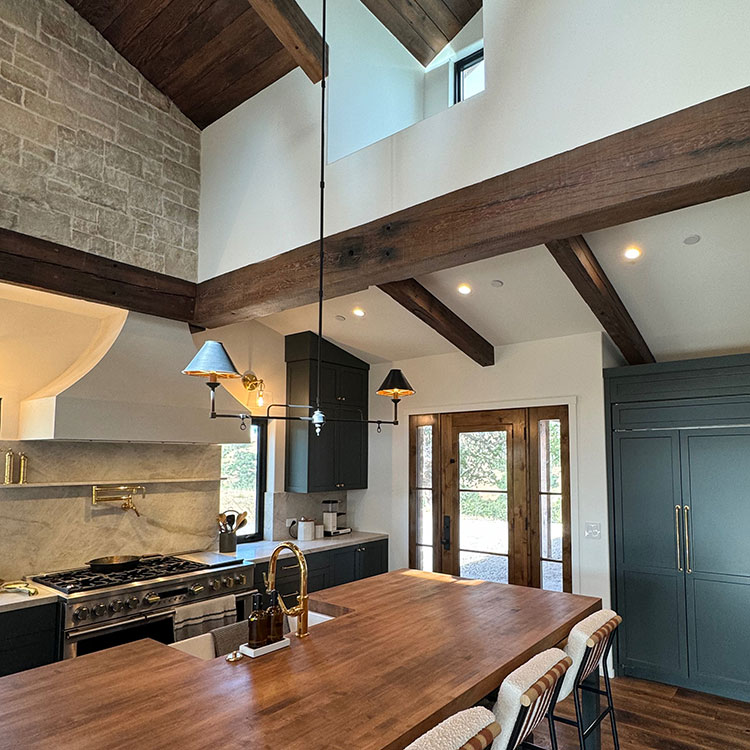 Modern kitchen interior with wooden ceiling beams, large wooden island countertop, dark cabinetry, a gas stove, and pendant lights above.