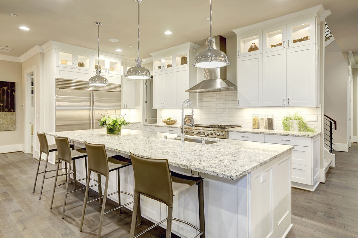 Spacious modern kitchen with white cabinetry, large granite island with four brown bar stools, stainless steel appliances, and pendant lights.