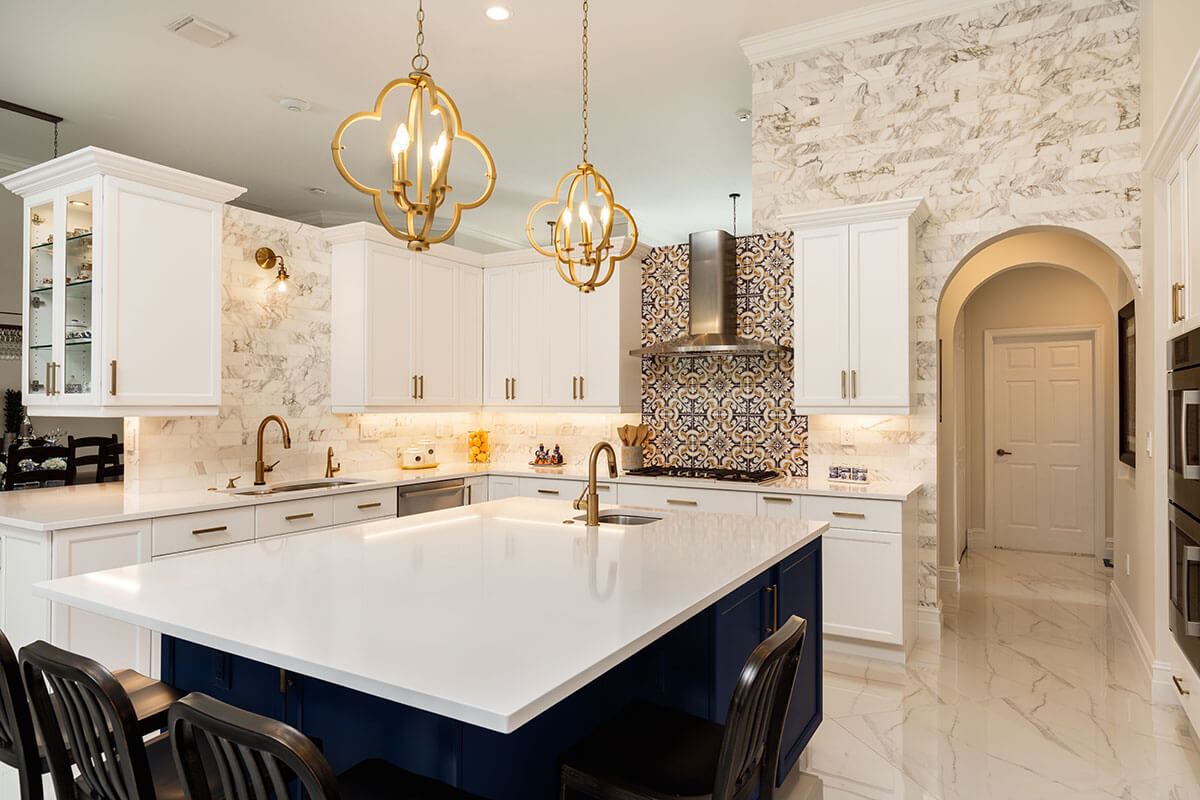 Modern kitchen with white cabinets, dark blue island with white countertop, gold fixtures, and decorative tile backsplash behind the stove.