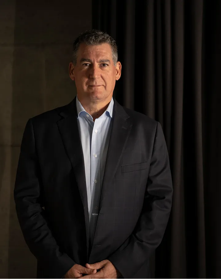 Man in a dark blazer and light blue shirt standing against a dimly lit dark background, looking forward with hands clasped.