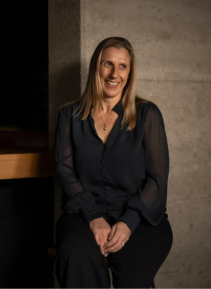 Woman with long blonde hair wearing a dark blouse and necklace, sitting and smiling against a concrete wall background.