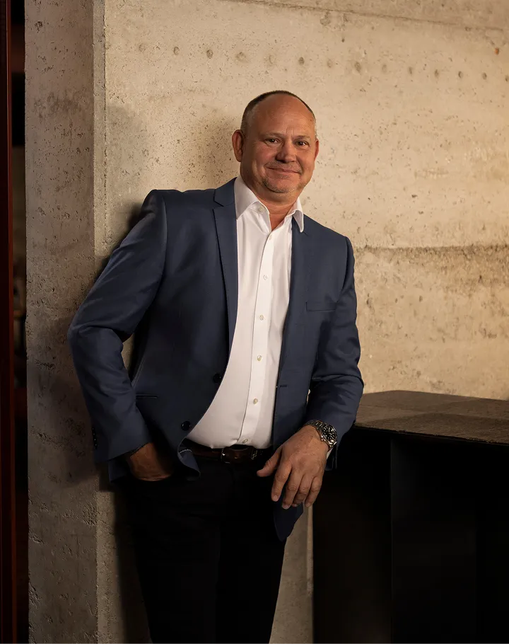 Man in a blue blazer and white shirt leaning against a textured concrete wall, smiling with one hand in his pocket.