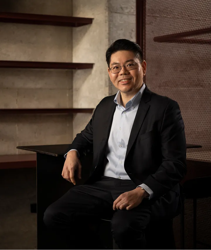 Man wearing glasses and a dark suit sitting on a chair in a room with empty wooden shelves on the wall.